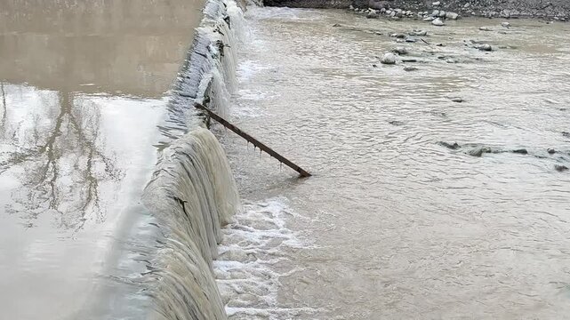 Water flowing over small dam edge, strong stream cascade into river, urban runoff scene, dynamic water movement, outdoor environment concept