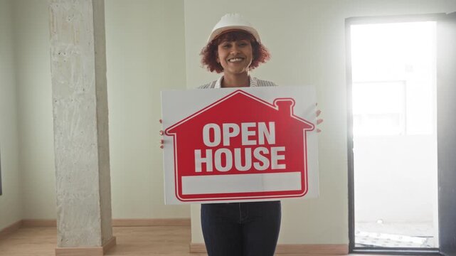 Woman holding open house sign, wearing hardhat and smiling inside the empty building; welcoming sale.