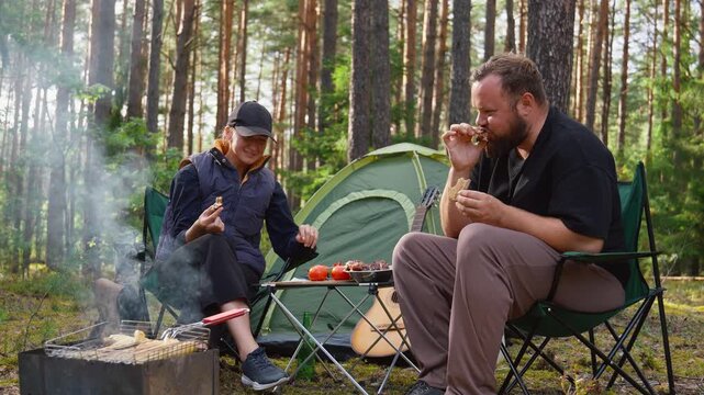 Young couple enjoying a picnic while sitting on chairs in a beautiful pine forest, with the woman slicing bread and the man taking vegetables from the table next to their tent and barbecue