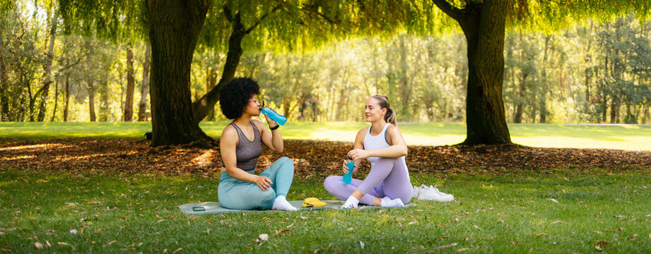 Two healthy women resting and hydrating in a park after outdoor fitness