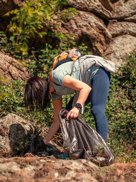 Mature female volunteer cleaning up garbage and plastic waste in a forest. She is wearing a backpack and gloves, collecting litter to protect the environment during an organized cleanup action.