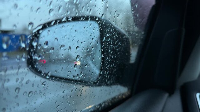 Close-up of a car side mirror covered in heavy raindrops. The reflection shows a wet road, moving vehicles, and a gloomy overcast sky.