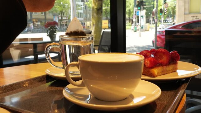 A cozy cafe scene featuring a clear glass mug with a tea bag steeping in hot water and a white ceramic cup of cappuccino on a tray. A strawberry tart is partially visible. The setting is by a window, 