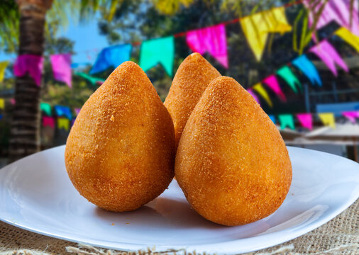 Three delicious Brazilian chicken coxinhas on a white plate, set against a vibrant Festa Junina background with colorful flags. Perfect golden-brown crust and traditional teardrop shape.