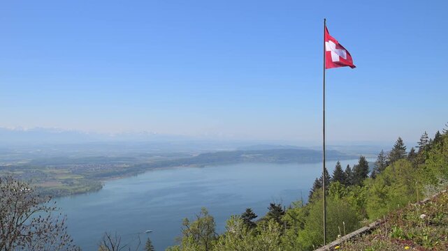 Scenic viewpoint with Swiss flagpole above Lake Geneva, forested hillside, and distant Alps under clear blue sky.