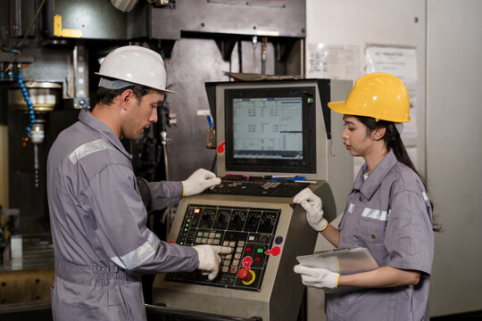 Adult male operator adjusts CNC controls while Asian woman technician checks settings with tablet, supporting machine monitoring, production accuracy, and teamwork in smart factory workflow daily