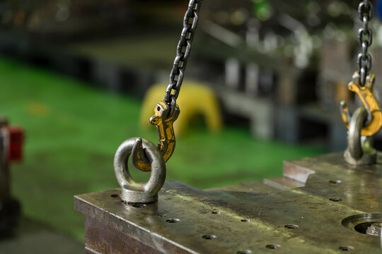 Yellow chain hook locks into steel lifting eye above heavy die block, supporting controlled hoist connection, safe mold transfer, and maintenance preparation in modern manufacturing workshop