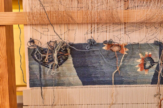 A close-up view of a wooden weaving loom with a partially completed tapestry in progress. Vertical warp threads are stretched taut across the wooden frame, with a section of woven textile visible