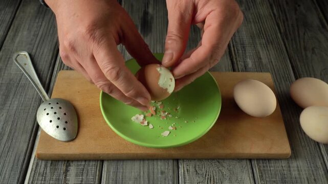 Hands carefully break eggshell and peel egg over green bowl on wooden cutting board, pieces of shell fall inside, other eggs and spoon present in kitchen