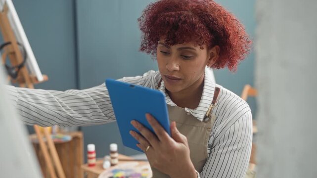 Woman holding blue tablet and reaching to canvas in studio, wearing apron and striped shirt; concentration.