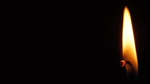 Macro shot of a burning candle flame positioned on the right side of a black background. Glowing candle wick flickering in the dark with large copy space.