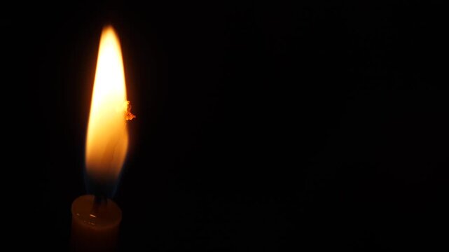 Burning candle on the left side of the frame with copy space on black background. Bright single candle flame close-up in complete darkness