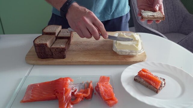 man making sandwiches with butter and salmon front view, kitchen food preparation process with slicing bread and spreading butter, preparing snack meal, close-up action assembling fish sandwich