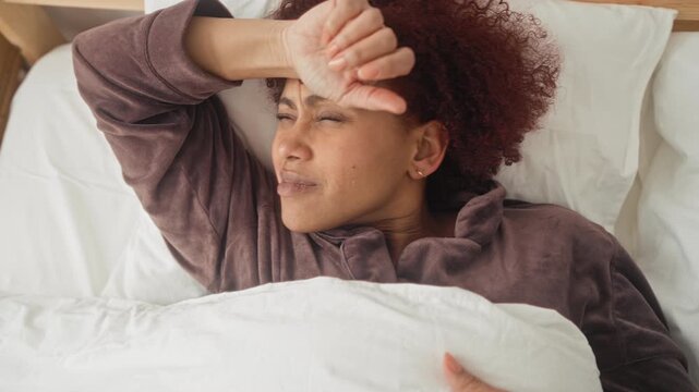 Woman in bed shields eyes with hand and raises arm over pillow and duvet, squinting and wincing in a building; morning tiredness.