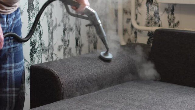 A person uses a professional handheld steam cleaner to deep clean and sanitize a fabric sofa in a home interior