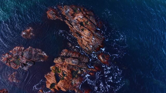 Aerial top view of coastal rocks and blue water. High angle top-down shot of weathered brown rocks in clear blue sea water with small waves.