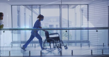 Naklejka premium Walking nurse wearing blue scrubs and mask pushing wheelchair along clinic hall with glass railing