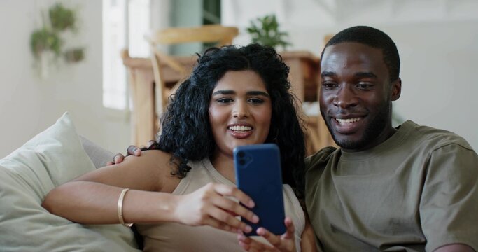 Smiling couple sitting on light sofa at home, woman wearing beige top holding blue smartphone