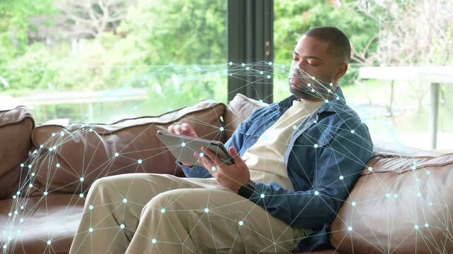 Man tapping tablet, scrolling and rotating while network overlay around sofa showing data for tech