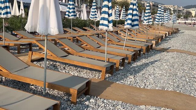 Beach Chairs and Umbrellas: A pristine beachside lounge area is set up with numerous wooden deck chairs and striped umbrellas, ready for visitors to relax under the sun. Nice, France
