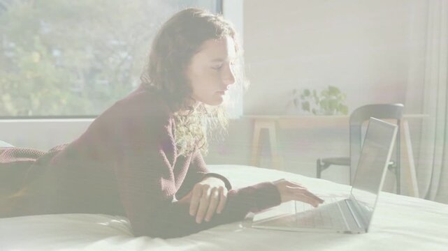 Woman lying bed by window, typing, smiling as ping causing animated overlay on laptop for studying