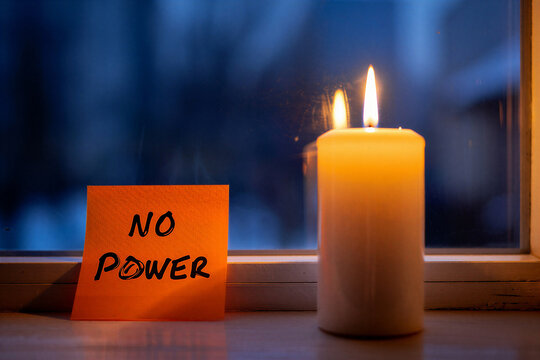 A burning white pillar candle illuminating an orange "NO POWER" sticky note by a window.