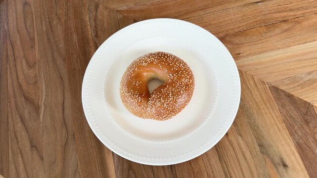 Video of a hand placing a sesame seed bagel onto a white plate on a wooden table, simple food preparation and breakfast concept.