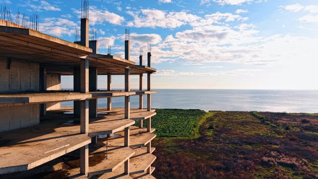 Unfinished multi story concrete building frame near sea coast. Exterior view of abandoned concrete construction site with pillars and floors overlooking the ocean and green field.