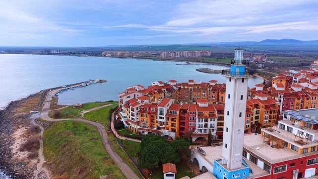 Aerial view of white lighthouse and coastal resort buildings. Drone shot of a lighthouse tower next to orange roofed apartment complex by the sea shore.