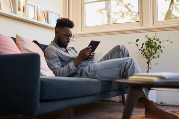 Fototapeta premium Man using tablet reclining on sofa in living room during daytime