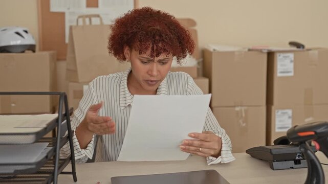 Woman holding document, finger to temple at desk inside building filled with cardboard boxes; small business stress.