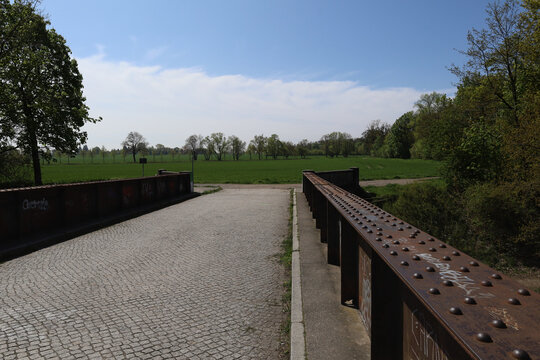 Historische Stahlbr&uuml;cke mit Kopfsteinpflaster &uuml;ber den Saalekanal, Blick auf gr&uuml;ne Wiesen und Fr&uuml;hlingslandschaft bei Sonnenschein, Burghausen, Leipzig, Sachsen, Deutschland