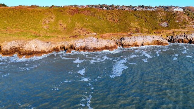 High angle shot of rocky cliff face and sea. Vertical aerial view of steep coastal cliffs and turquoise ocean water with white foam in Wales