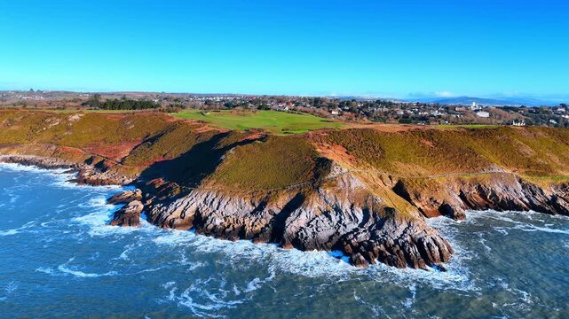 High angle view of rocky headland and coastal path Wales. Aerial shot of jagged rocks green fields and residential area along the Welsh coastline near Gower peninsula
