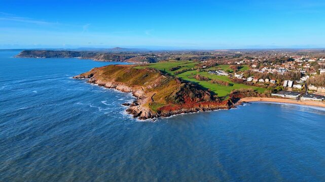Panoramic aerial shot of Gower Peninsula coastline and golf course. Drone perspective of rocky headland, green fields, golf course and coastal town under clear blue sky in Wales.