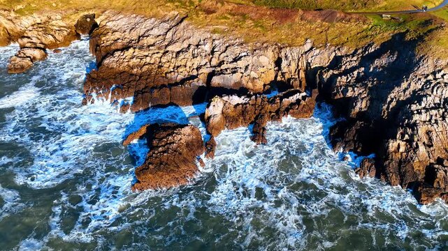 Top down view of white sea foam hitting rocky cliffs. Close-up aerial shot of crashing ocean waves on textured rocks with high contrast shadows and golden sunlight.