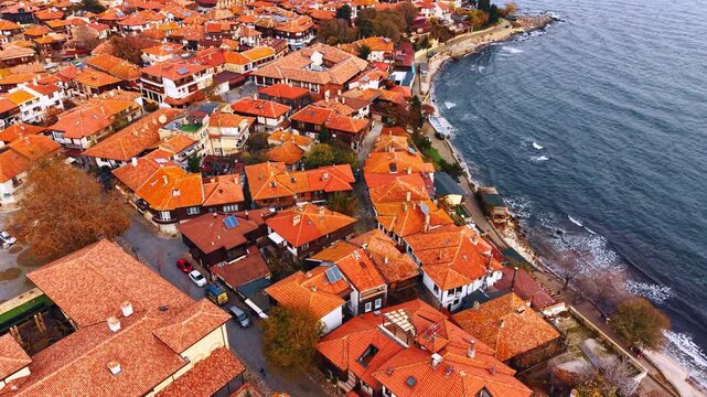 Moving over the tiled red roofs of the houses in the dense urban landscape along the shore. Picturesque coast of Nesebar, Bulgaria. Aerial view.