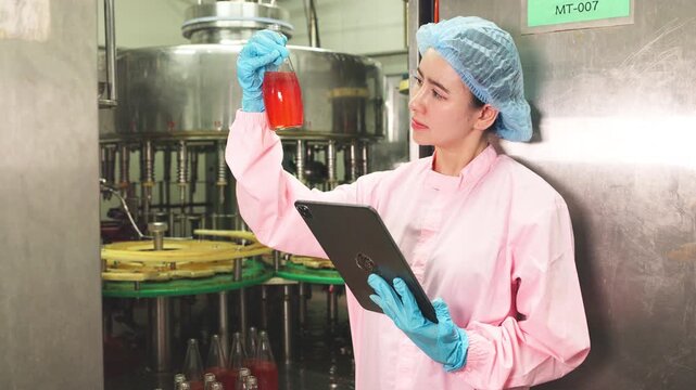 Female factory worker in protective clothing examining liquid sample in glass bottle while recording data for quality control in production facility