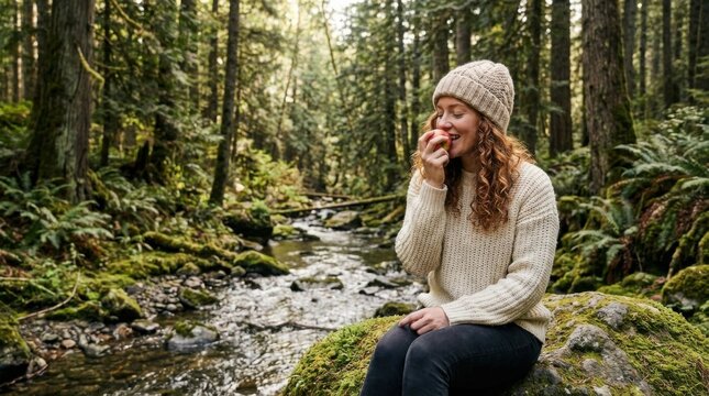 Curly haired woman wearing a cream sweater and knitted beanie eating a red  by a rocky stream in a damp temperate forest.