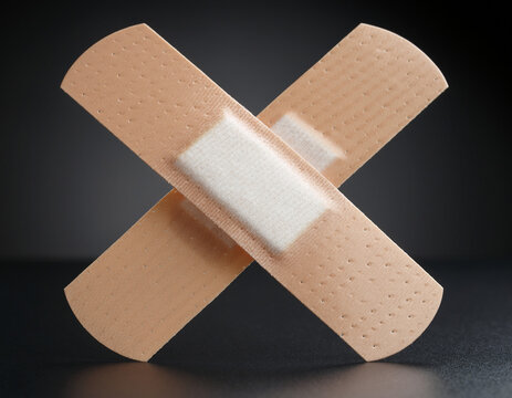 Two adhesive bandages are placed in an X formation on a dark background. These medical plasters are for wound care and are part of first aid supplies for cuts and scrapes. They symbolize the need for 