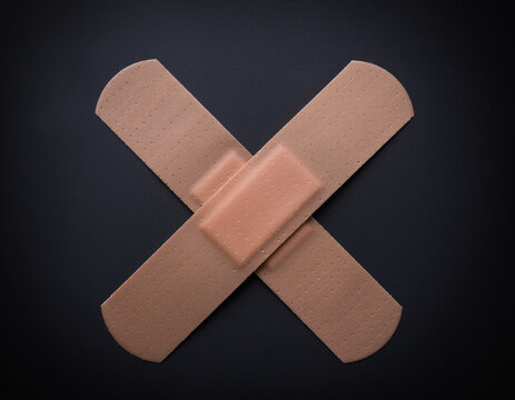 Two adhesive bandages are placed in an X formation on a dark background. These medical plasters are for wound care and are part of first aid supplies for cuts and scrapes. They symbolize the need for 
