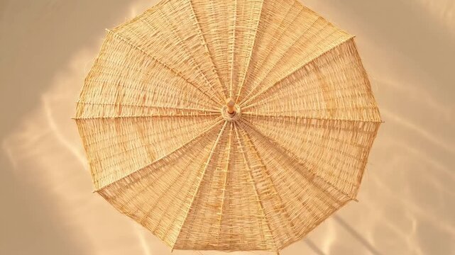 Overhead view of a natural woven straw parasol backlit by bright sunlight, casting intricate shadows and creating a warm golden glow against a neutral background.
