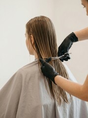 Naklejka premium Female hairdresser in black gloves cuts long hair of client wearing gray cape in salon
