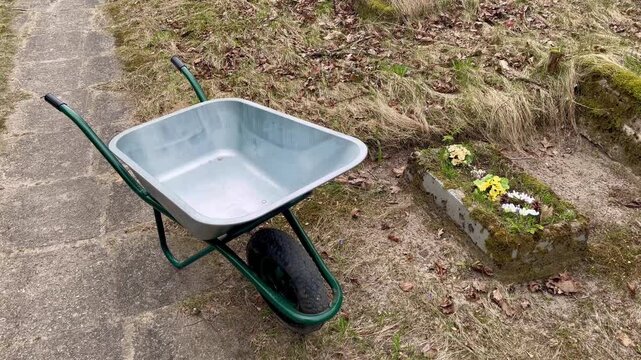 Empty wheelbarrow standing near small grave with flowers in cemetery during spring maintenance showing preparation for cleaning and care of burial site