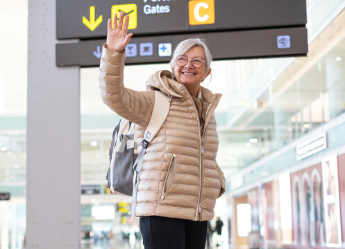 Smiling senior woman waves goodbye in a bright airport terminal wearing a puffer jacket and backpack as she prepares for travel. Friendly, confident traveler enjoying the journey and departure process