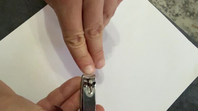 Macro shot of person clipping nails with a metal nail clipper on a white surface. Clean grooming concept, personal hygiene and everyday self-care at home