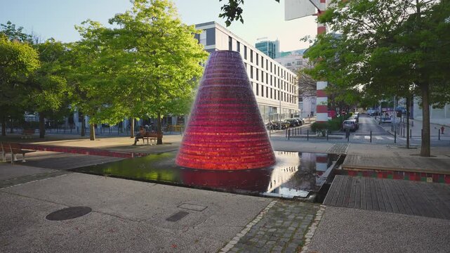 LISBON, PORTUGAL - JANUARY 15, 2026: Red and pink conical mosaic fountain active in a modern city square surrounded by trees and pedestrian area