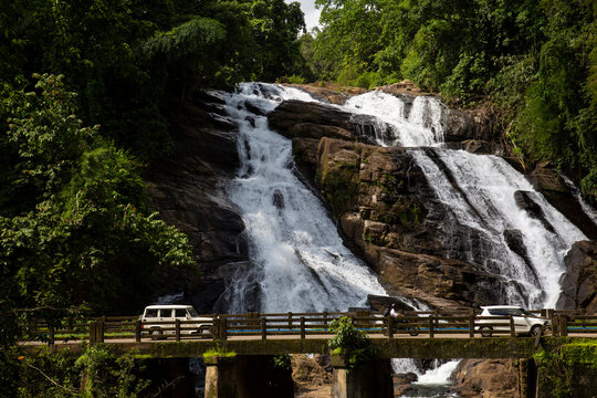 Charpa waterfall cascading over rocky terrain near Athirappally, Chalakudy, Kerala, India. Road bridge with passing vehicles in foreground amid lush tropical rainforest.
