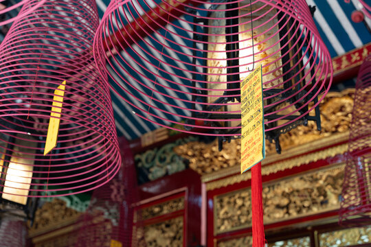 Pink spiral incense coils hang from the ceiling of a Chinese assembly hall in Hoi An, Vietnam, with yellow prayer tags and a richly gilded red lacquer altar glowing in the background.