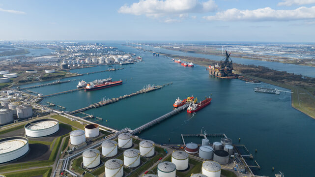 Europoort Rotterdam, Netherlands - 15 March 2025: Aerial view of Europoort Rotterdam industrial port with oil storage tanks, cargo ships docked at piers and wind turbines under a blue sky.
